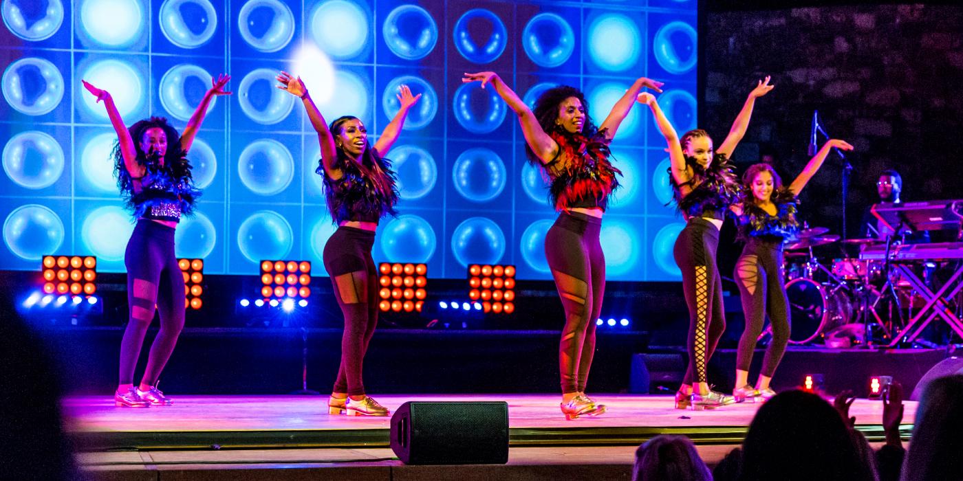 Five women dance on a stage with blue lights in the background