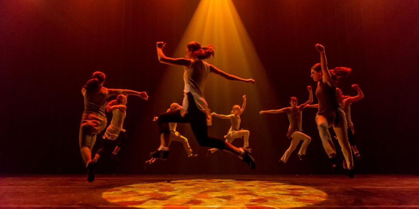 Trinity Irish Dance Company members leaping on stage in a circle with an orange spotlight shining down on them