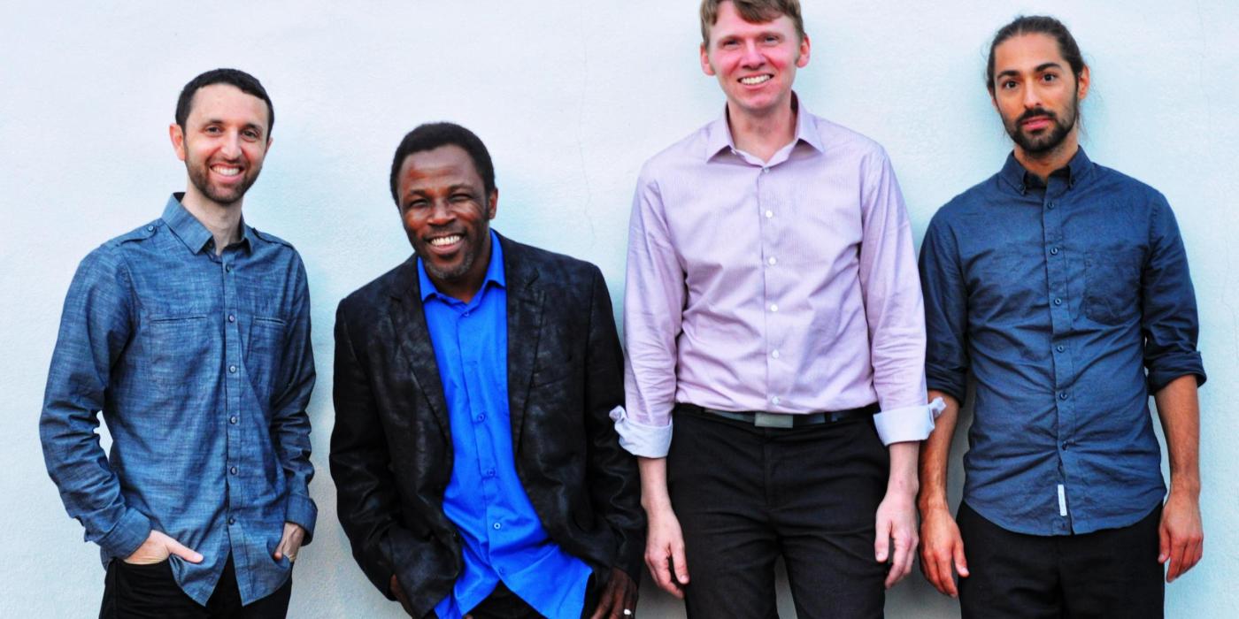 Four men stand against a white wall smiling at the camera. Three wear blue button-up shirts and one wears a pink button-upshirt. 
