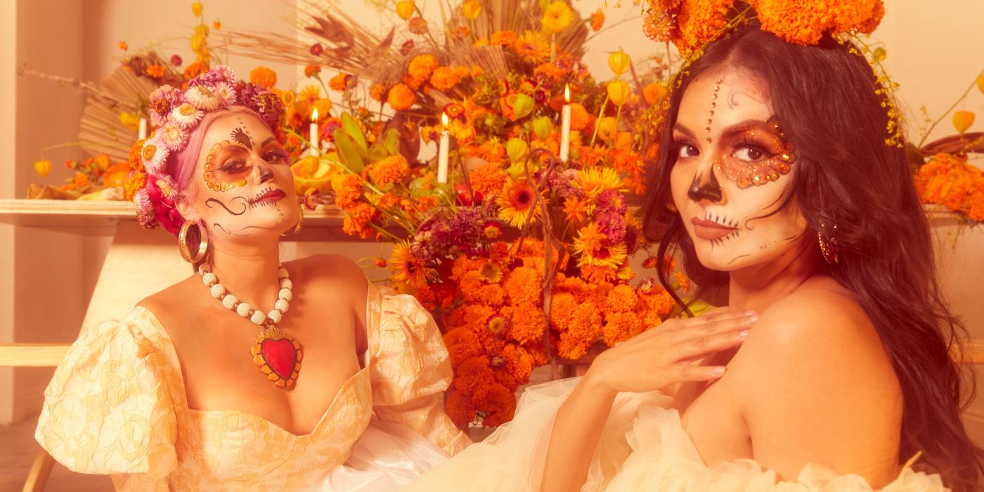 Two women pose in white dresses, orange flowers behind them and their faces painted like skeletons