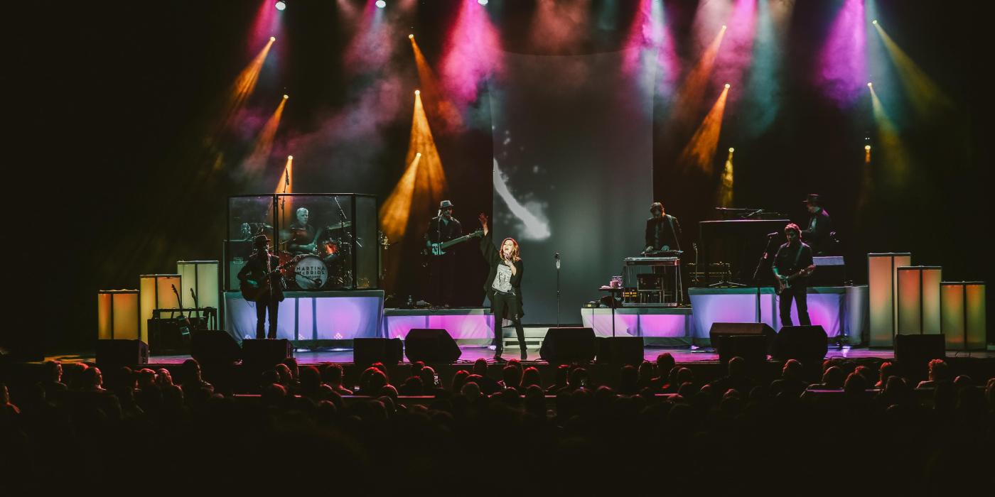 A woman dressed in black raises her hand to the sky as she sings on a stage filled with colorful lights and a crowd watches