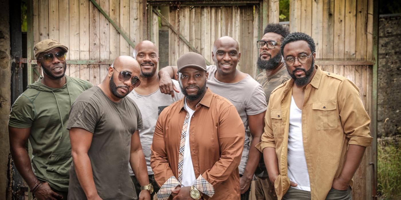 The seven male singers of Naturally 7 pose in front of a building, wearing clothes in natural color tones