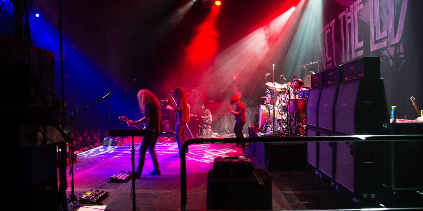 The image is a back stage view of a rock band playing with colorful lights overhead - a guitarist and drummer are in the foreground