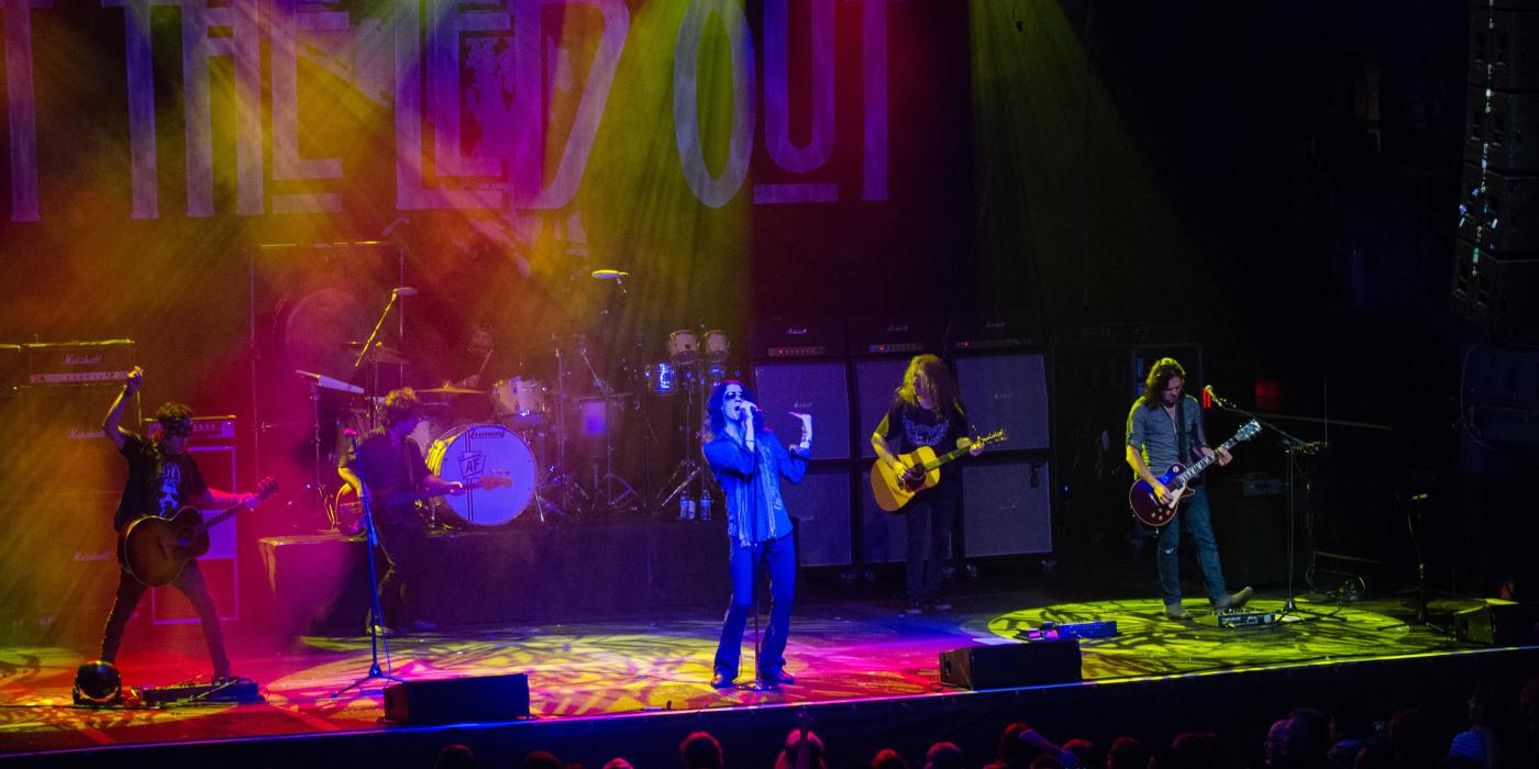 Wide shot of a stage with pink and yellow lights overhead, three guitarists, a singer and drummer are on stage