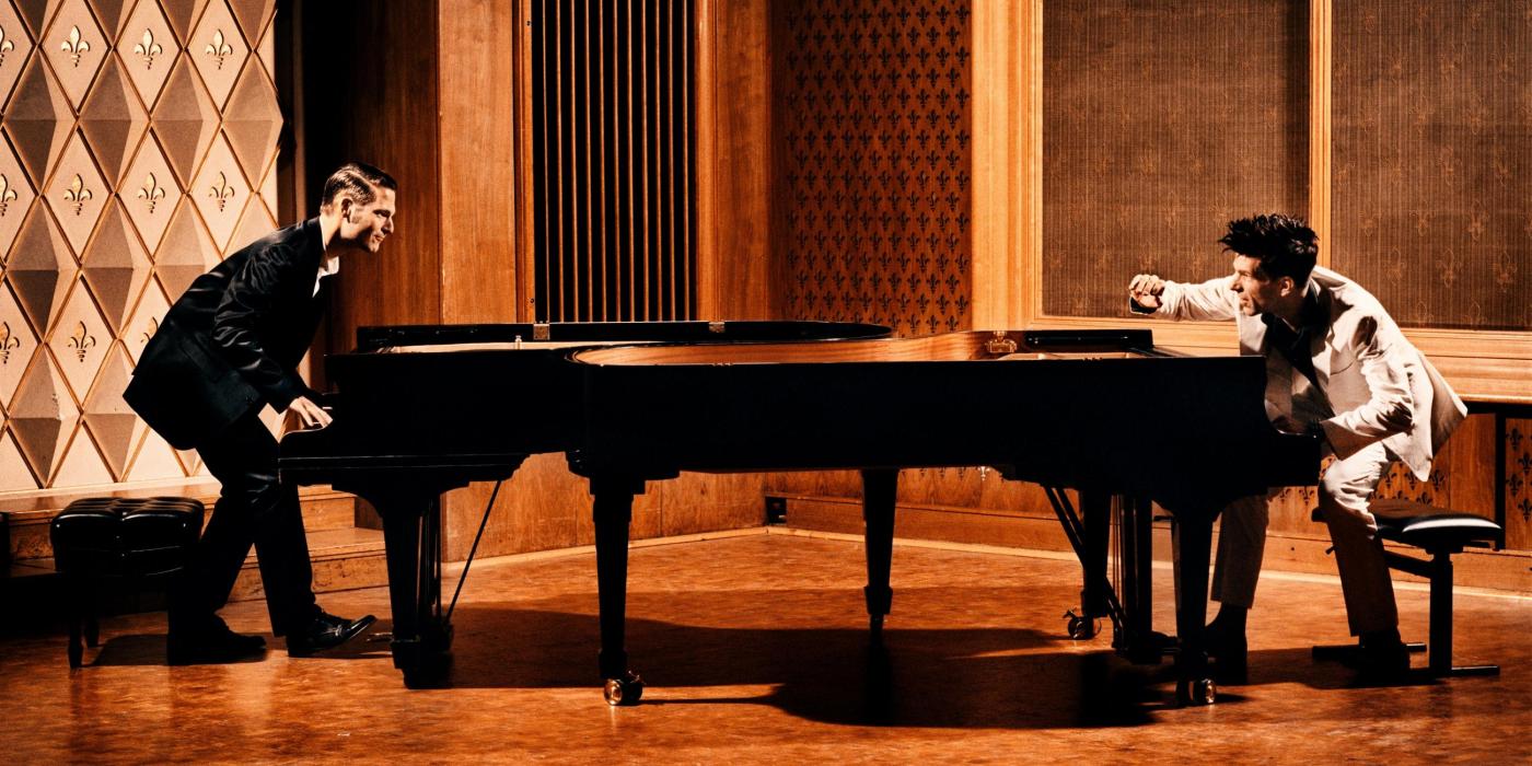 Two pianists, one dressed in white and one in black, face each other while playing a piano.