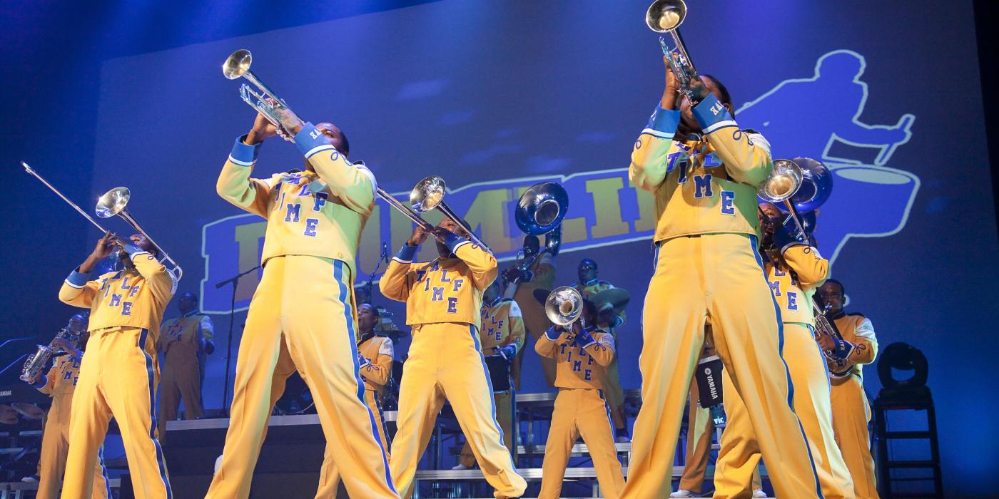 Horn line from Drumline Live wearing yellow uniforms in front of a blue backdrop.