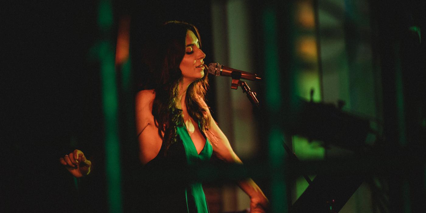 A woman with long brown hair wears a green dress, eyes closed as she sings into a microphone