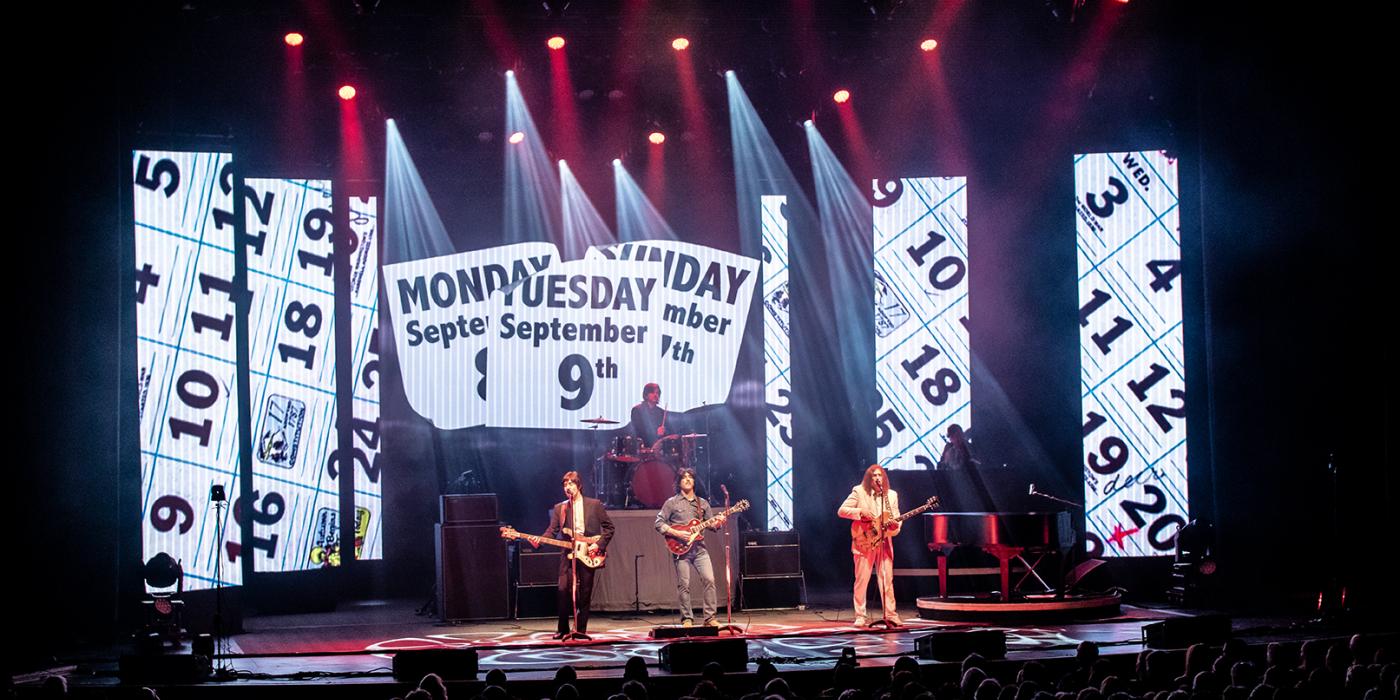 Three men dressed in clothes from the 60s stand on a stage with screens and lights behind them