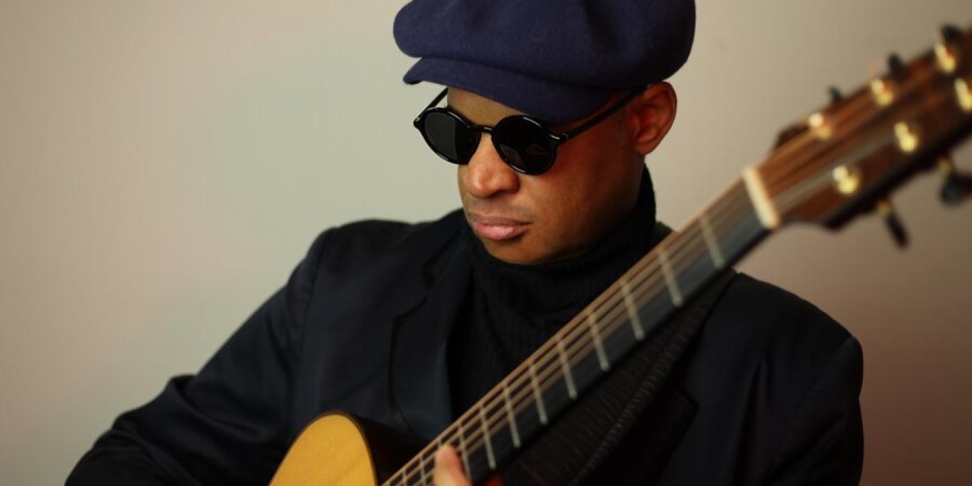 A man holds a gold colored acoustic guitar with his fingers on the fret. He wears all black with dark glasses, a turtle neck, jacket and drivers hat.