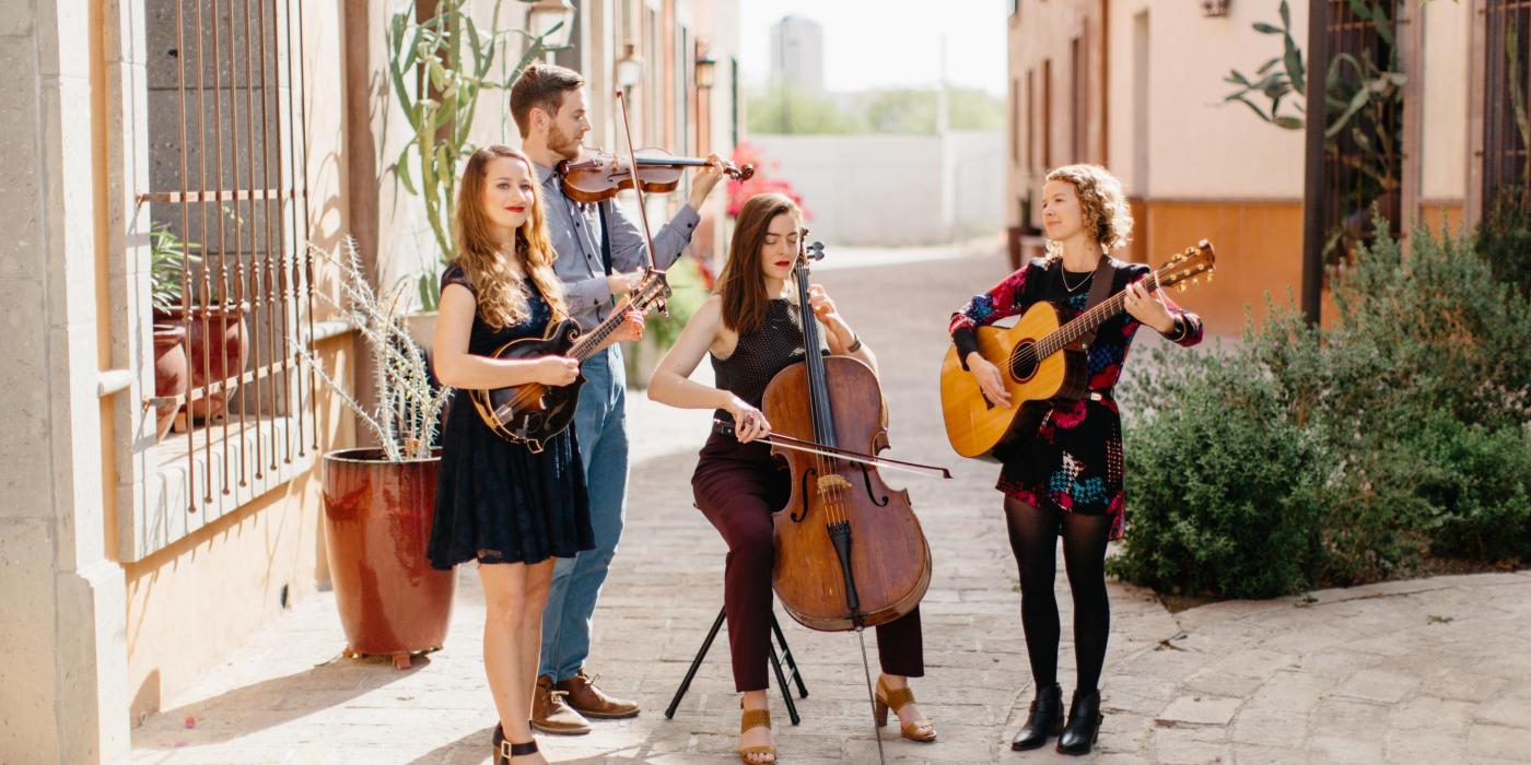 Three women and a man play their string instruments and sing in a sunny cobblestone square between adobe buildings.