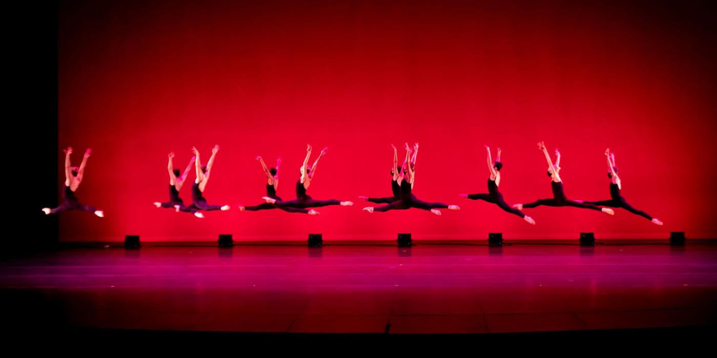 A row of ballet dancers wearing black costumes leap in front of a red background.