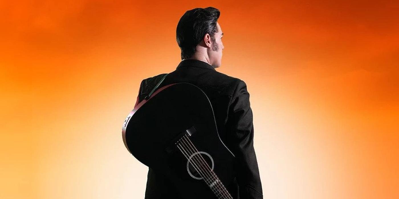 A man in black has his back to us, standing on railroad tracks with a guitar on his shoulder