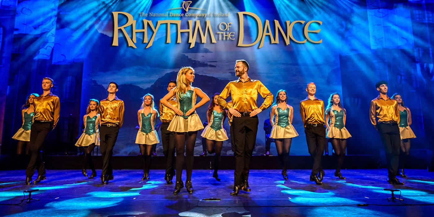 A troupe of Irish step dancers wear yellow shirts and black pants or skirts with blue lights behind them.