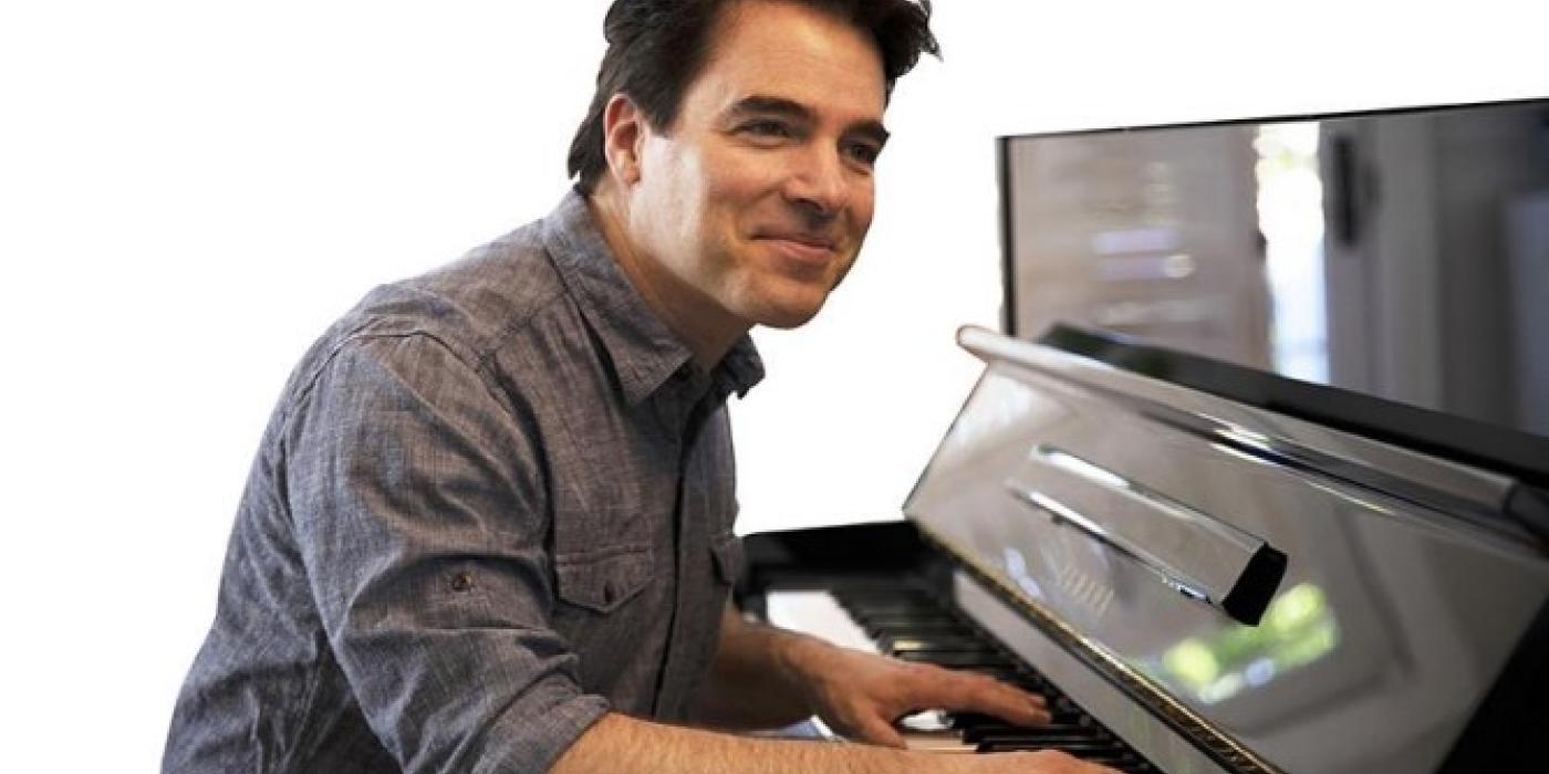 A wearing a grey shirt looks at the camera smiling, while playing the piano