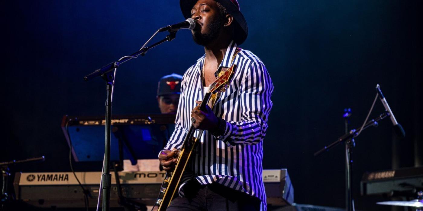 A man stands on a stage behind a microphone, singing, eyes closed. He is wearing a black and white striped button up shirt and a brown fedora. A guitar is slung low on his hip as he strums. 