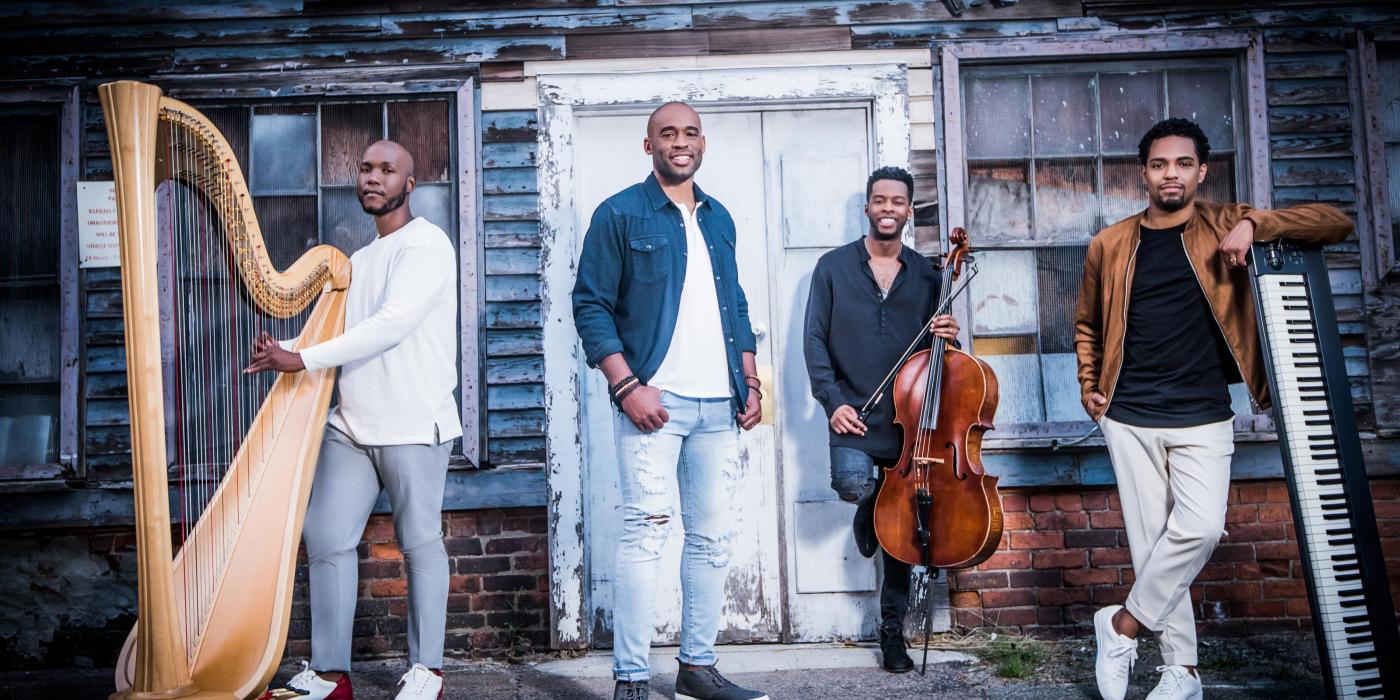 Four men stand casually in front of a weathered gray house with a white door. The main singer stands in the middle with the others gathered around with their instruments - a cello, harp and keyboard