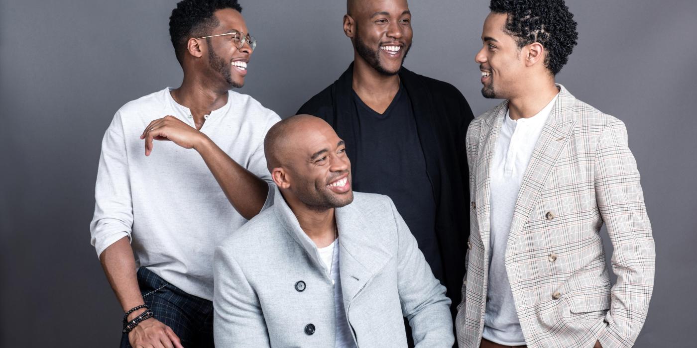 Four men pose for a formal photo with a gray background. All of them are smiling and laughing at one another, wearing gray, light blue and black. 