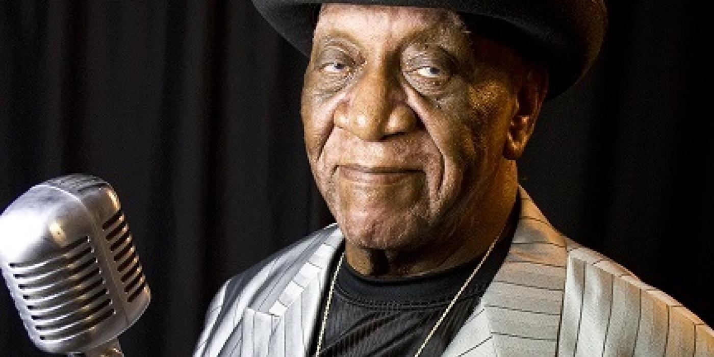 Big Pete Pearson wearing a silver suit and black bowler hat standing behind an old style microphone