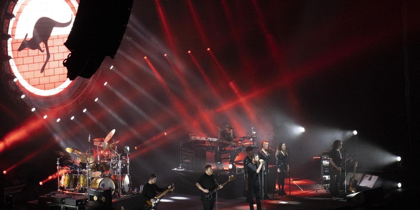 A band stands on a stage with red laser lights and an LED screen projecting a kangaroo