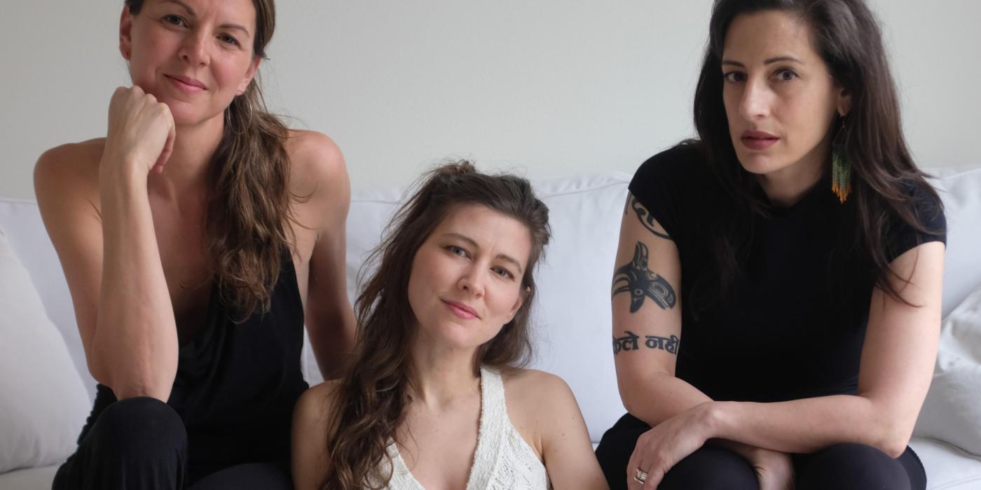 Three young women look forward at the camera with seriousness, two with brown hair and one with blond hair. 