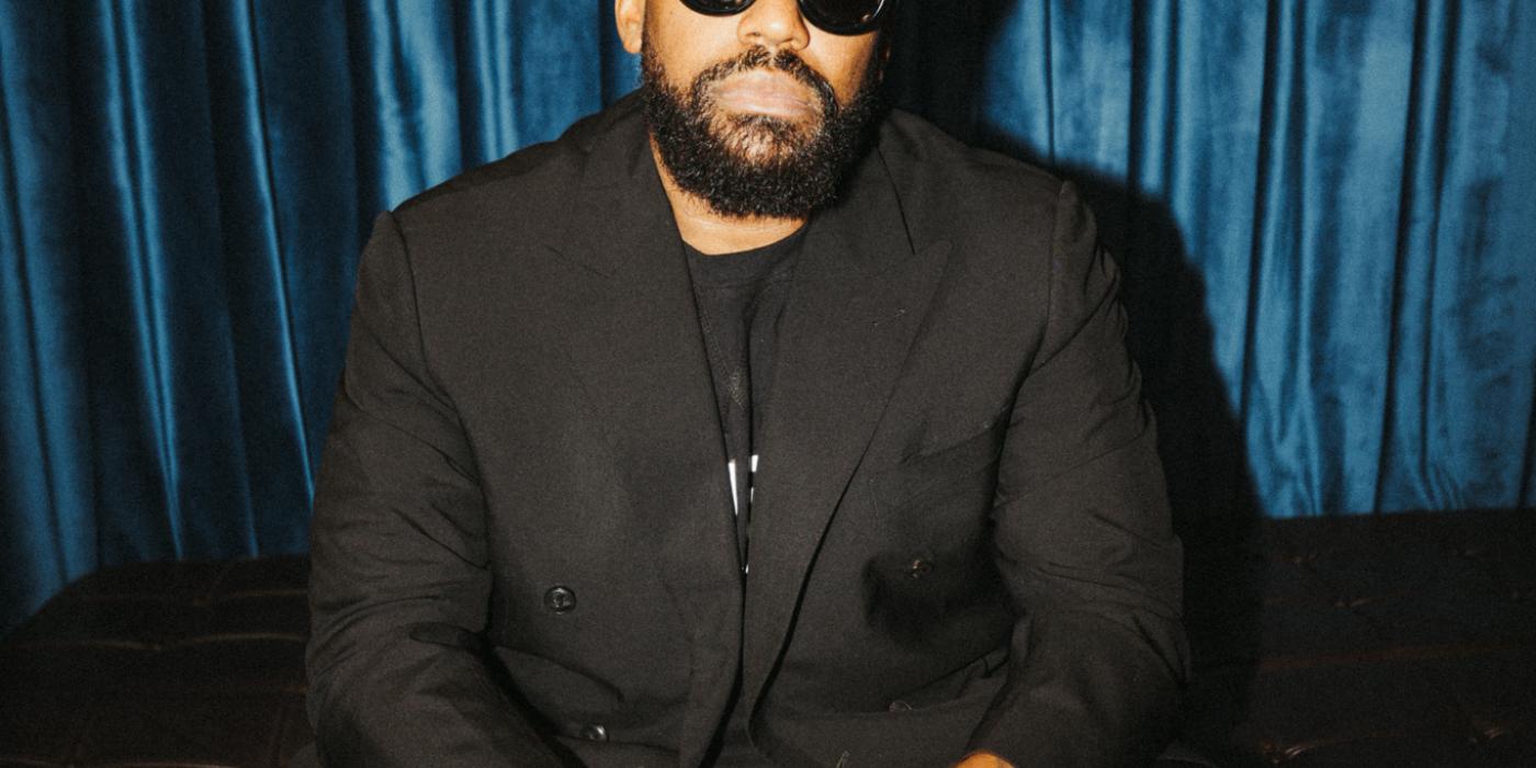 A man wears a black shirt, black suit jacket, black beanie hat and black sunglasses, facing the camera 