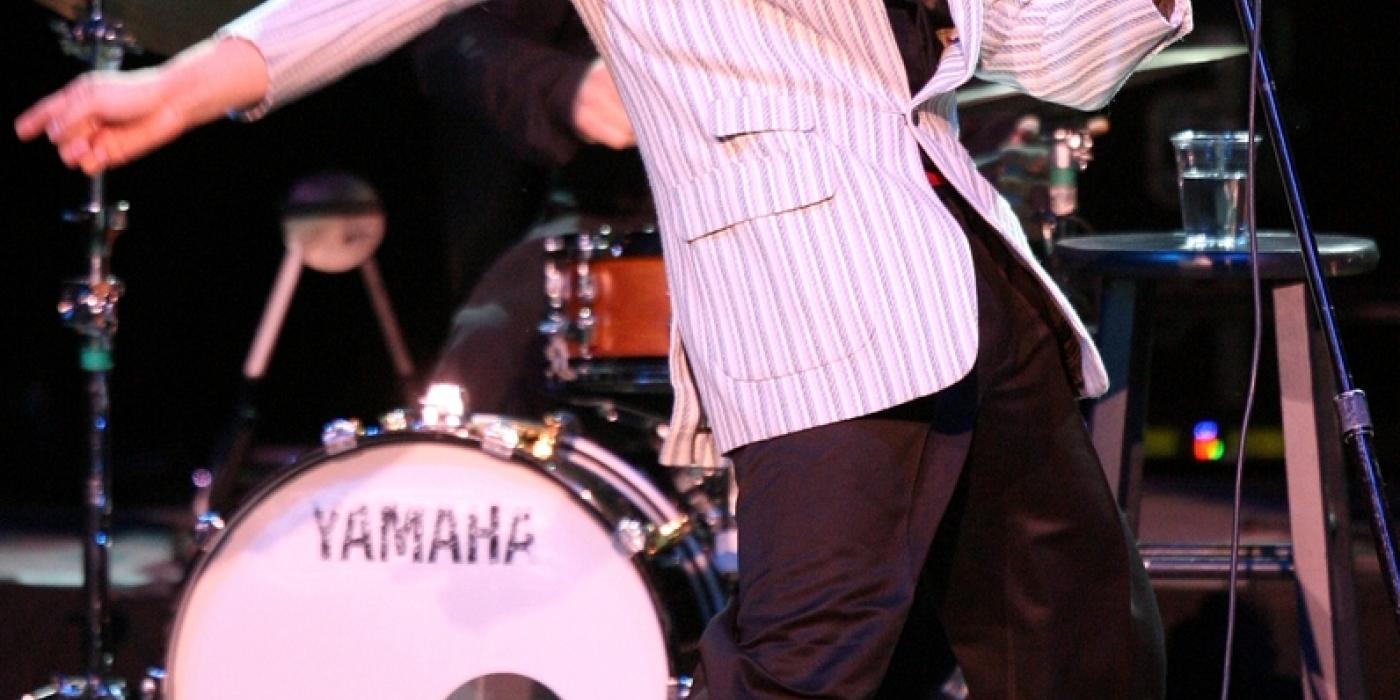 Man in a striped white suit and black pants swaying his hips to the side while holding a microphone