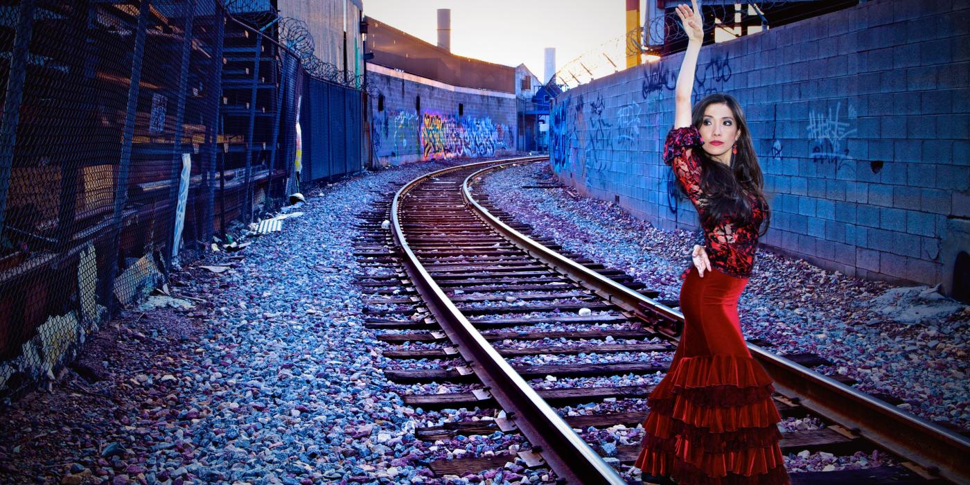 A tall woman poses by railroad tracks in a red velvet dress with layers of ruffles from her knees to the ground