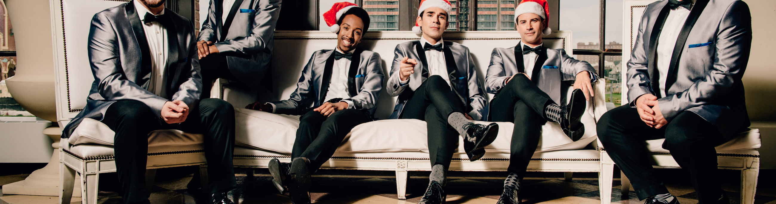 Six men dressed in black and gray suits and wearing santa hats, sitting on a couch looking at the camera