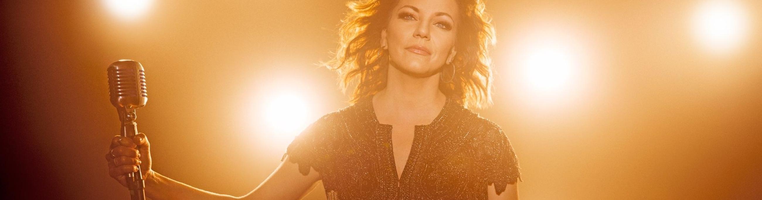 woman with brown hair looking at the camera holding a microphone. There are bright lights all around her
