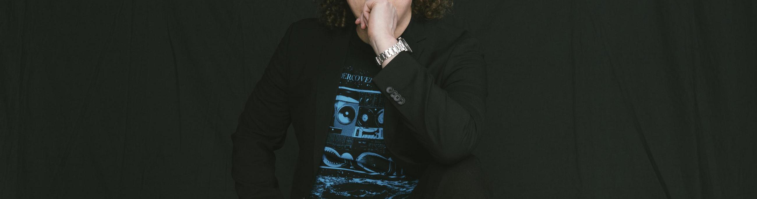 white man with curly brown hair wearing all black and a black hat in front of a black background.