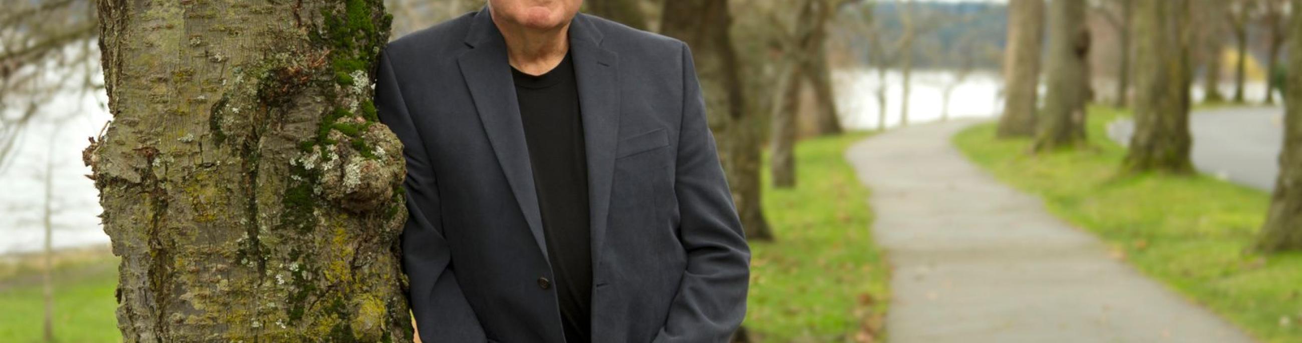 Paul Green poses outdoors, leaning on a tree.