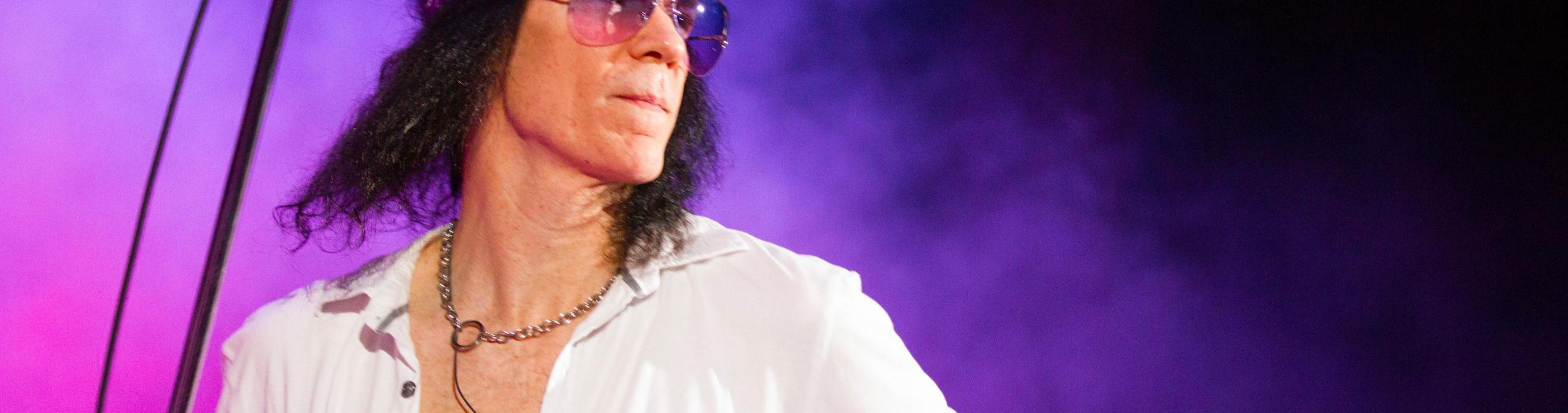 Paul Sinclair has shoulder length black hair, wearing a white button up shirt open at the chest and sunglasses.