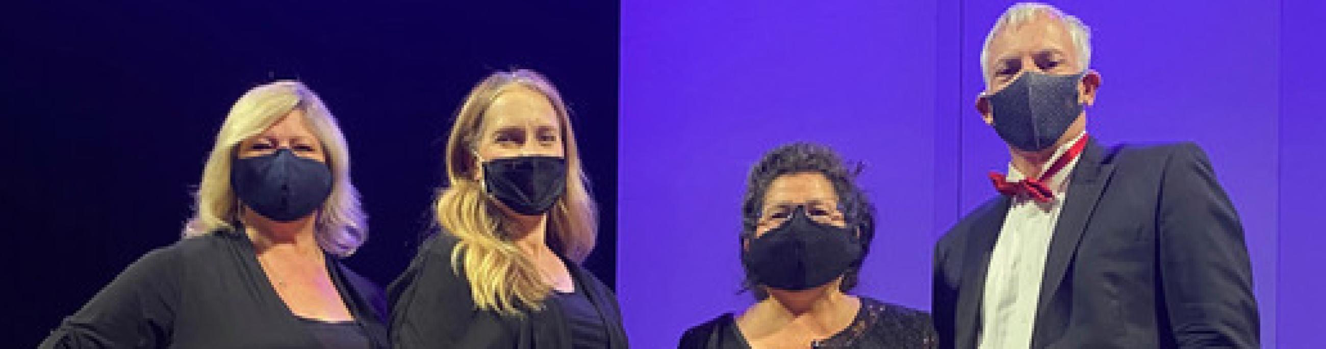 Four musicians, dressed in formal wear and masks, pose in front of a purple backdrop.