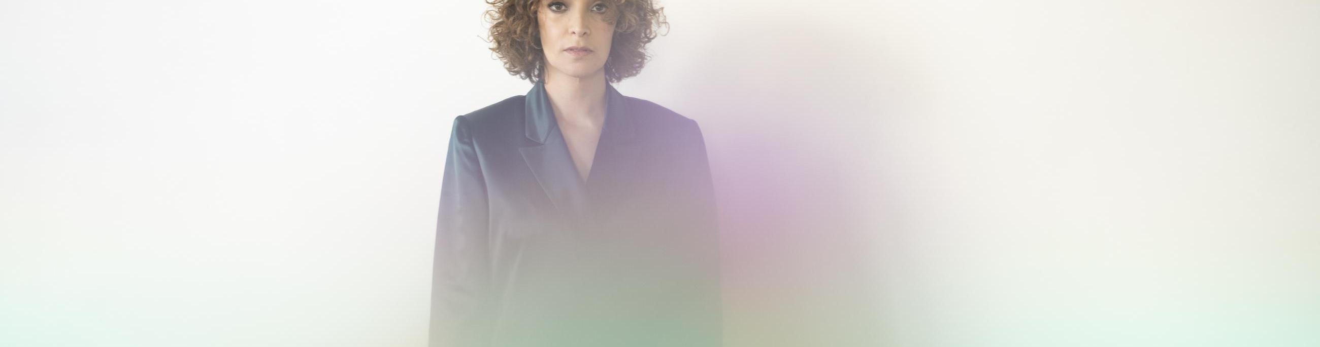 Woman with lighter skin and brown curly hair standing in the center, looking at the viewer. There is a rainbow like fog in front
