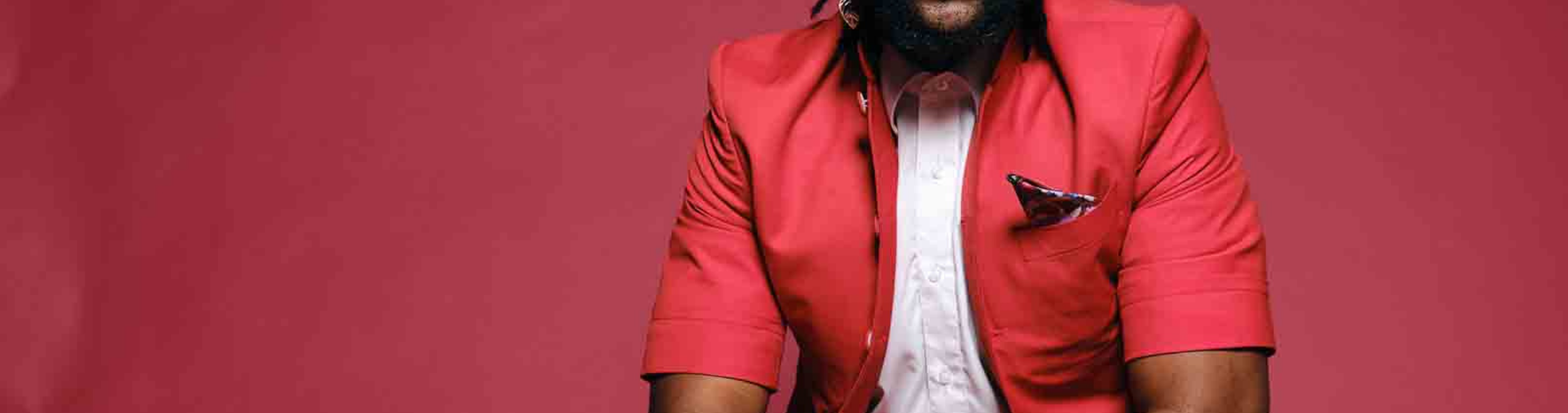 Michael Mwenso, a man with long dreadlocks in a ponytail, sits in front of a red wall in a red suit