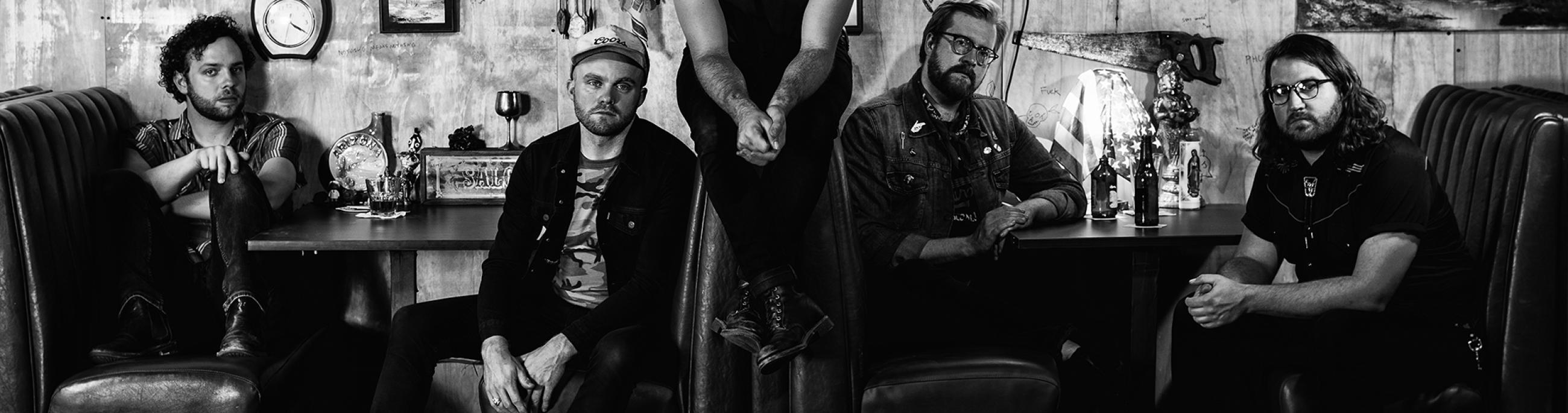 In a black-and-white photo, five male band members of Jared and The Mill sit in a booth at a diner.