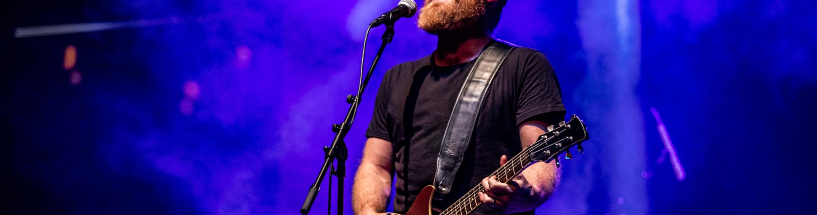 A man with a beard and black drivers cap, sings into a microphone and plays guitar