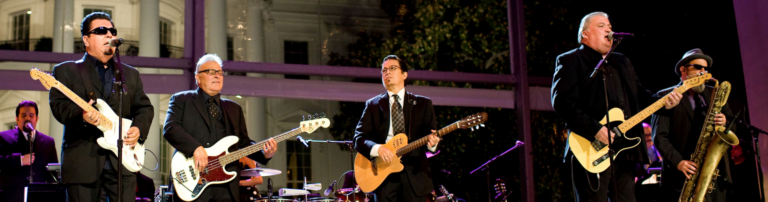 four men on stage playing instruments and singing, dressed all in black