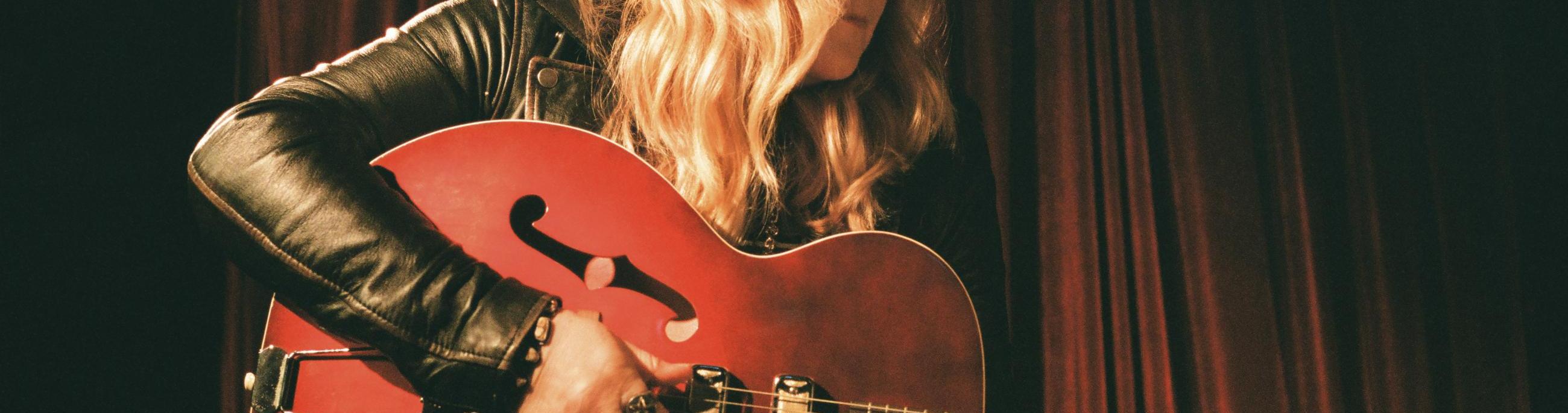 Blonde woman holding a red guitar looking to the right with a dark background