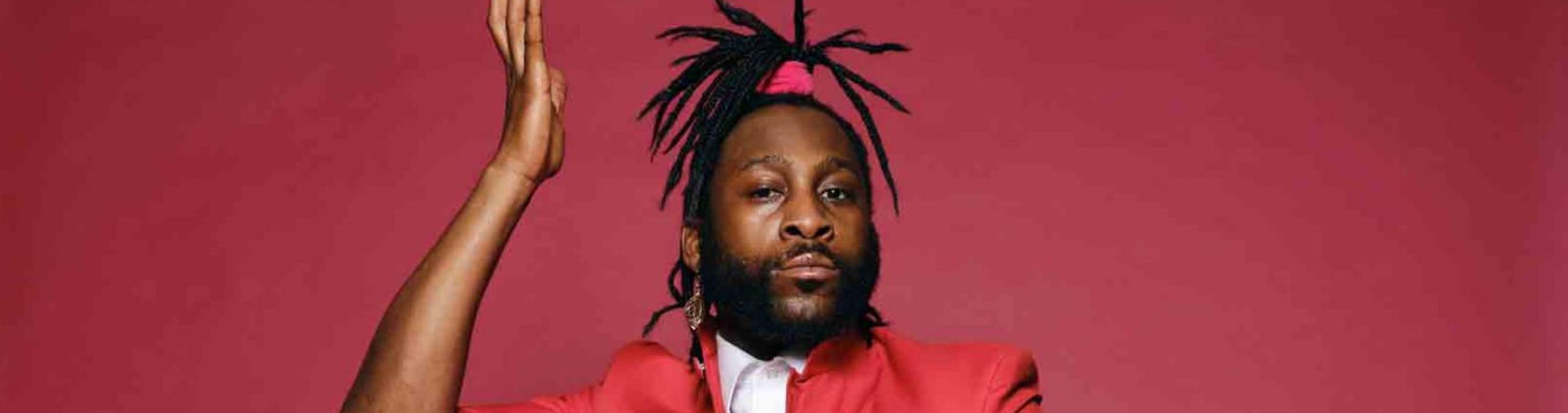 A man with dreadlocks in a ponytail wears a red suit, sitting on a chair in front of a red background. He looks at the camera.