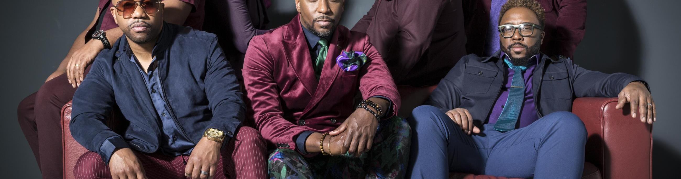 seven black men sit on a red leather couch looking at the camera