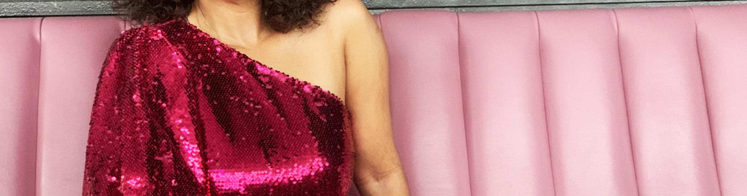 A woman with dark curly hair sites on a pink leather couch, wearing a magenta sequined top