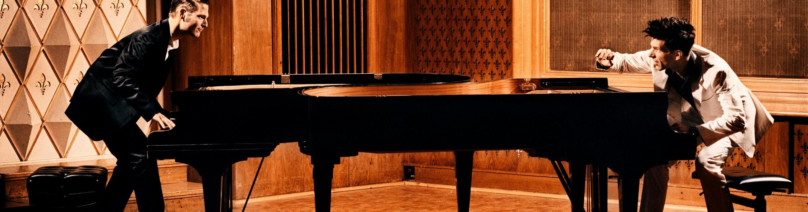 Two pianists, one dressed in white and one in black, face each other while playing a piano.