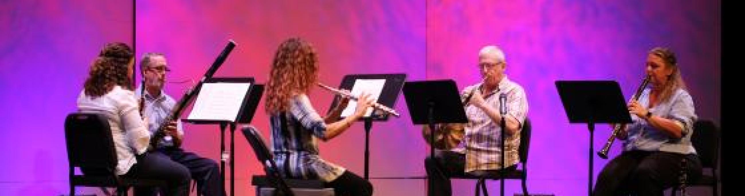 A wind quintet performs in front of a pink and purple backdrop.