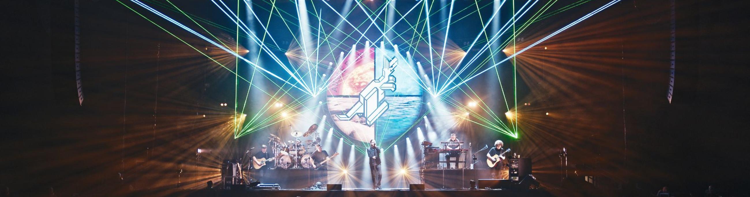 Stage with a lot of light lines shooting upward. A band on the stage and a crowd of people in front of them