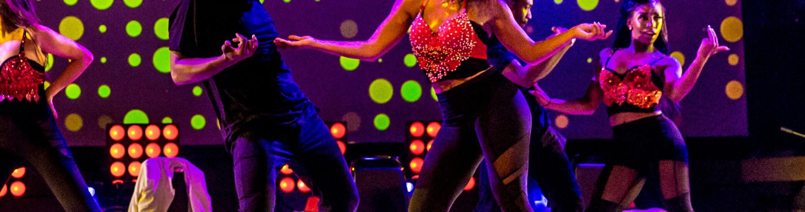 3 women and one man tap dancing, looking at each other. The lights are a purple/green and they are all wearing black and red