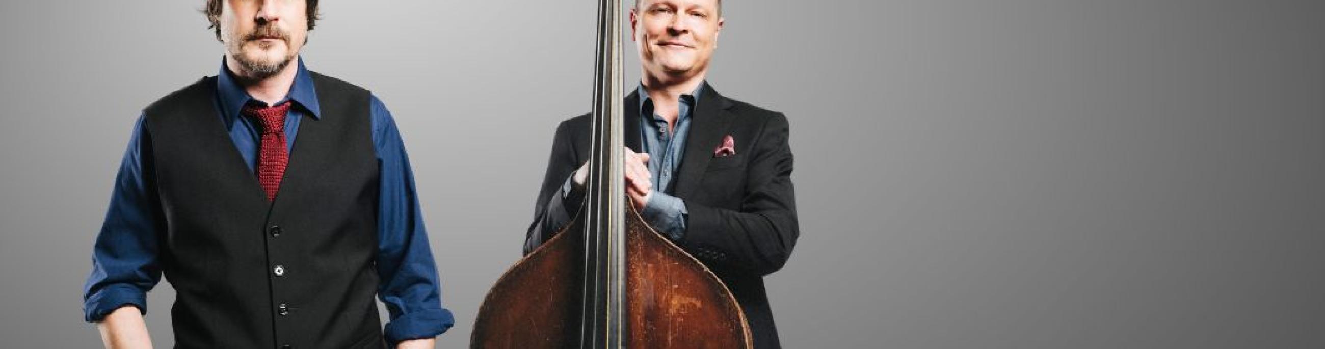 two men standing next to each other, both white, one wearing a hat and the other holding a bass