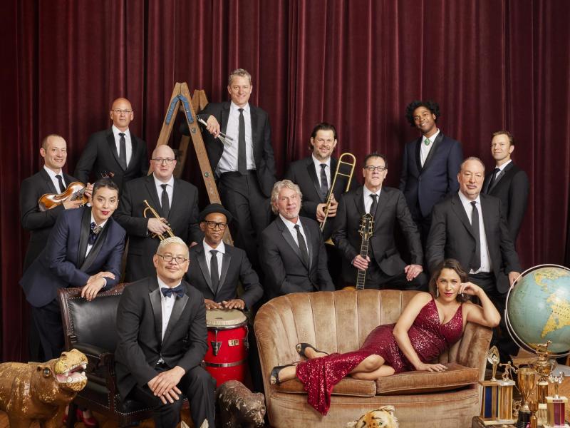 Thirteen men wearing black suites and a women in a red dress laying on her side on a sofa. 