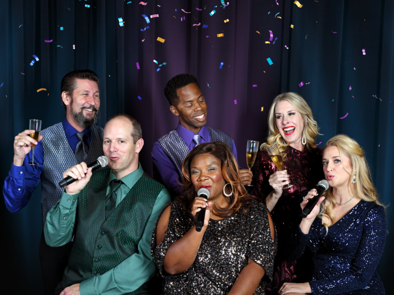 Group of adults with 3 singing into mics and other 3 holding champagne flutes