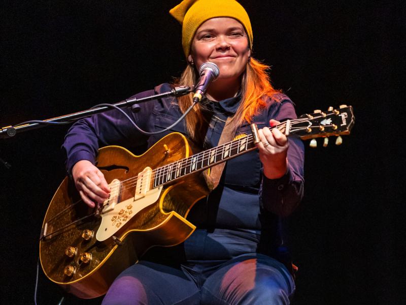 Young tan woman wearing a yellow beanie, playing a guitar on stage.